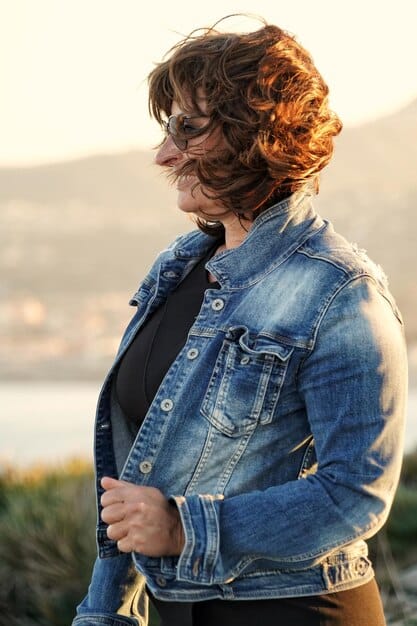 A man with long, flowing hair styled in a loose wave, wearing a casual denim jacket, standing against a natural outdoor backdrop, epitomizing a relaxed and stylish vibe.