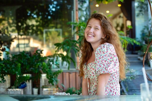 A woman with a round face smiling and showcasing a long bob with side-swept bangs. The background is an outdoor café setting.