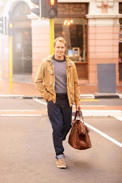 A man walking down a city street, carrying a stylish leather messenger bag over his shoulder. He is wearing a casual but put-together outfit, including jeans, a button-down shirt, and a light jacket.