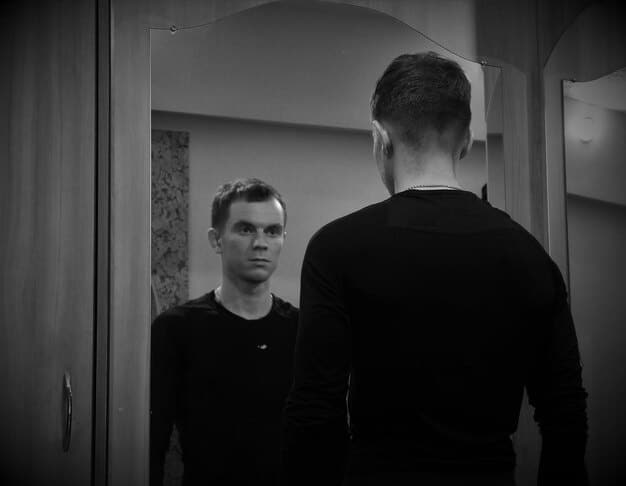 A black and white photograph of a man staring into a mirror, with a blurred reflection showing two faces, one confident and one sorrowful, representing the internal struggle between projected strength and hidden vulnerability.