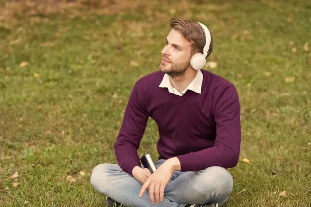 A man practicing mindfulness meditation outdoors with noise-canceling headphones. He's sitting cross-legged on a yoga mat in a peaceful garden, eyes closed, and hands resting gently on his knees. The background is blurred with lush greenery. Focus on his posture and relaxed expression.