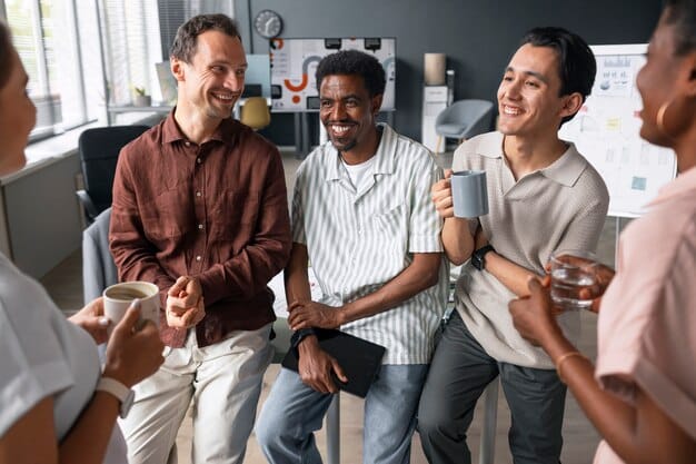 A group of male colleagues sitting together in a relaxed lounge area, engaged in conversation and smiling. The scene conveys a sense of camaraderie and mutual support among coworkers.