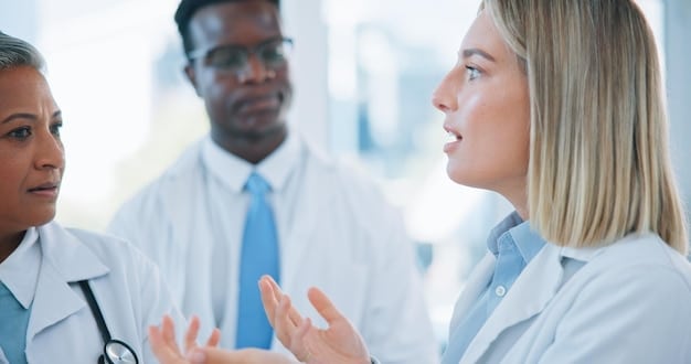 A close-up shot of a doctor in a white coat is having a conversation with a male patient while sitting at a desk in a well-lit clinic. The patient is a man in his late 50s or early 60s. The doctor is looking at the patient and explaining a concept.