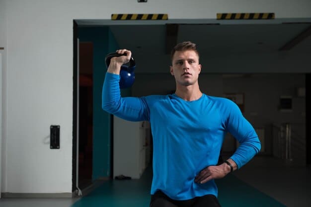 A photograph showing a man standing in the gym, taking a water bottle. The lighting from the gym makes a very good contrast with his body, and he is wearing athletic attire.
