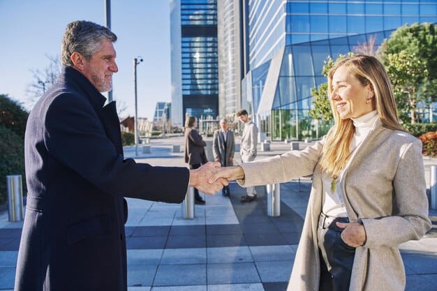 A person's hand extending to shake another person's hand, with a blurred backdrop of a bustling city street or office building. It symbolizes reaching out and connecting in a professional setting.