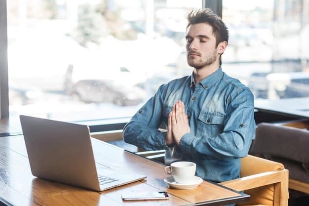 A man in a professional office setting, perhaps in front of a laptop, taking a moment to lightly press his palms together and close his eyes, indicating a brief mindfulness break during work.