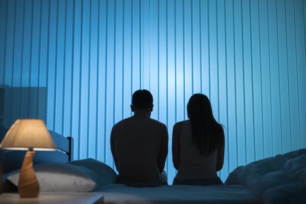A silhouette of a couple sitting far apart on a bed, facing away from each other, with a distinct blue, melancholic filter, symbolizing emotional distance and the impact of depression on intimacy.