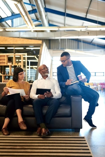 A diverse group of business executives are gathered in a modern office lounge, engaged in a lively discussion. Some are holding tablets and smartphones, presumably accessing LinkedIn. The scene suggests collaboration, networking, and the use of LinkedIn for professional connections and career advancement.