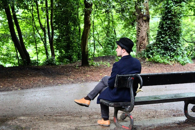 A single man sitting alone on a park bench, looking contemplative but not dejected. The angle is slightly from behind to show a sense of solitude without overwhelming sadness. The background includes other people enjoying the park, subtly highlighting the contrast between isolation and community, related to 