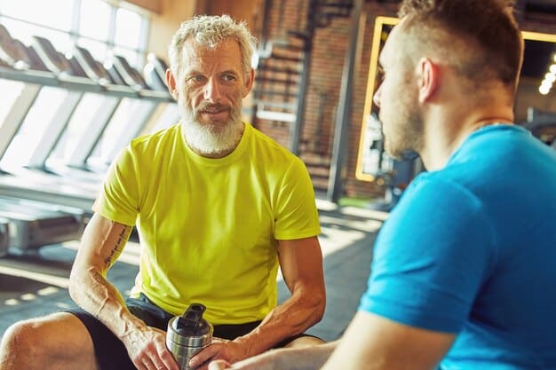A person in their early 40s is consulting with a personal trainer in a gym. They are discussing a workout plan while looking at a tablet. The setting is a modern gym with weights and machines, highlighting the expertise and guidance available for The Role of Exercise in Boosting Your Sex Drive: A Workout Plan for Enhanced Performance.